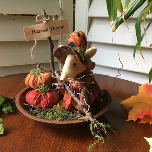May include: A whimsical autumn-themed centerpiece featuring a felt mouse wearing a hat and holding a twig broom. The arrangement includes fabric pumpkins, moss, and a sign that reads "Harvest Thyme". The display is set in a wooden bowl.