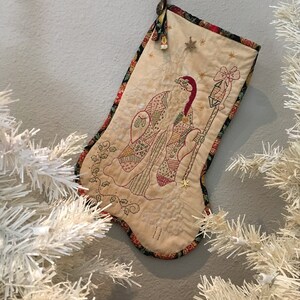 May include: A cream-colored Christmas stocking with an embroidered design of a figure holding a lantern. The stocking has a patterned border and is decorated with stars. It hangs from a loop with a button.