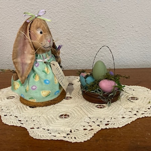 May include: A brown bunny figurine with long ears, wearing a mint green dress with yellow chick and pastel egg patterns. A tag reads "Happy Easter." A small basket holds colorful eggs and moss. The items sit on a cream-colored, lace doily.