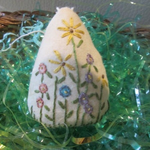 May include: A cream-colored, teardrop-shaped ornament embroidered with colorful flowers. The design features yellow daisies, pink and blue blossoms, and green stems. The ornament is set against a backdrop of green decorative filler.