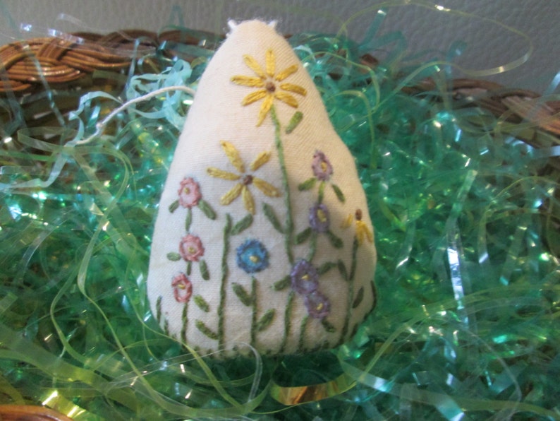 May include: A cream-colored, teardrop-shaped ornament embroidered with colorful flowers. The design features yellow daisies, pink and blue blossoms, and green stems. The ornament is set against a backdrop of green decorative filler.
