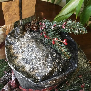 May include: A decorative winter-themed hat with a black brim and a snowy texture. The hat is adorned with pine needles, red berries, and pinecones. A small sign is attached to the hat. The hat is part of a holiday decoration.