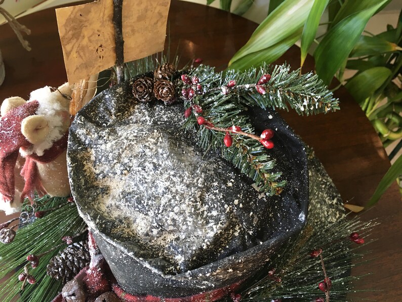 May include: A decorative winter-themed hat with a black brim and a snowy texture. The hat is adorned with pine needles, red berries, and pinecones. A small sign is attached to the hat. The hat is part of a holiday decoration.