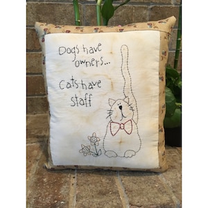 May include: Square cushion with a cream-coloured centre featuring a stitched cat design wearing a red bow tie. The cushion has the text "Dogs have owners... Cats have staff." The border is a tan fabric with a floral pattern.