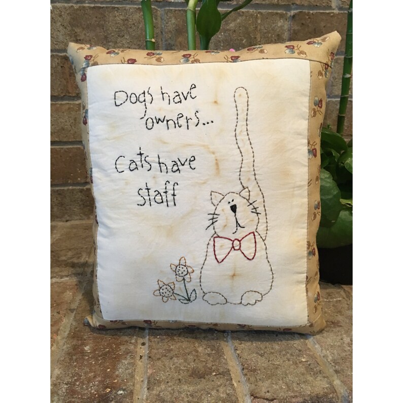 May include: Square cushion with a cream-coloured centre featuring a stitched cat design wearing a red bow tie. The cushion has the text "Dogs have owners... Cats have staff." The border is a tan fabric with a floral pattern.