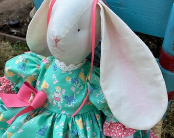 May include: A handmade white bunny doll with long ears and pink ribbon accents. The doll wears a teal dress with a pattern of children and animals, and a large pink bow. The doll's ears have pink inner linings.