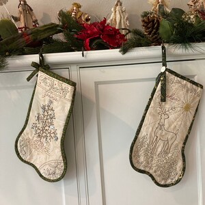 Puede incluir: Dos calcetines navide&ntilde;os color crema con ribete verde cuelgan de un armario blanco. Uno presenta un dise&ntilde;o de &aacute;rbol y el nombre "Jennie". El otro tiene un dise&ntilde;o de reno y el nombre "Cohen".