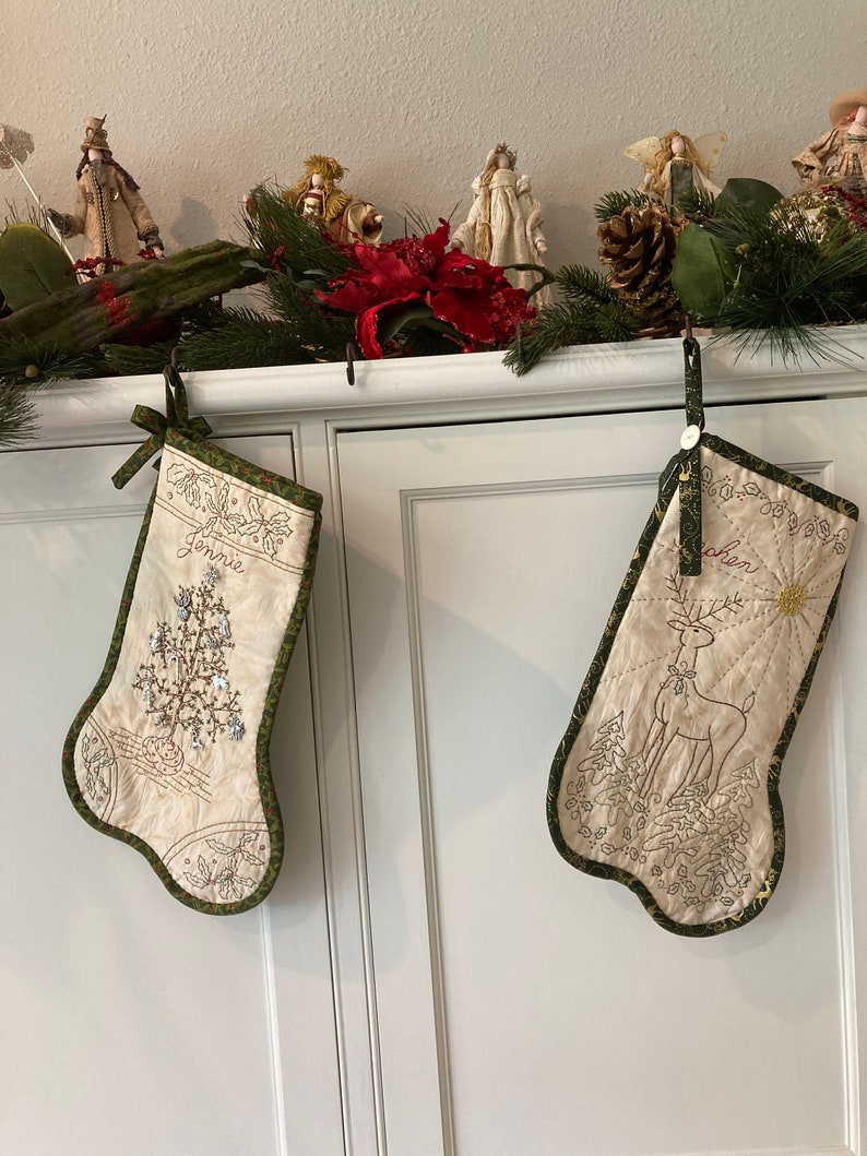 Puede incluir: Dos calcetines navide&ntilde;os color crema con ribete verde cuelgan de un armario blanco. Uno presenta un dise&ntilde;o de &aacute;rbol y el nombre "Jennie". El otro tiene un dise&ntilde;o de reno y el nombre "Cohen".