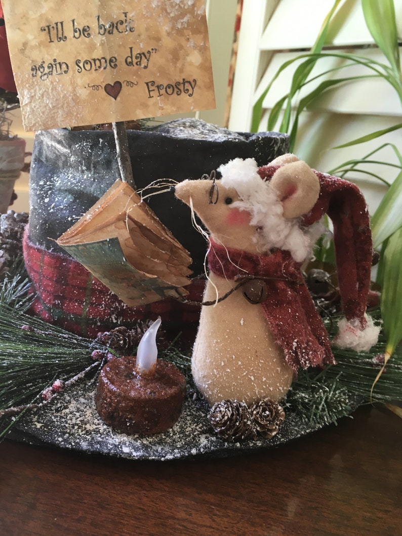 May include: A whimsical Christmas decoration featuring a mouse wearing a Santa hat and scarf, with a small candle and a sign that reads "I'll be back again some day" Frosty. The mouse is tan with pink ears and a red scarf.