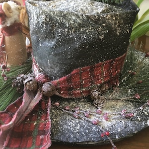 May include: A decorative black top hat with a red and black plaid ribbon, adorned with pine needles, berries, and small bells. The hat is dusted with a snowy effect, creating a festive holiday centerpiece.