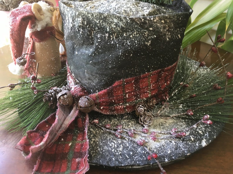 May include: A decorative black top hat with a red and black plaid ribbon, adorned with pine needles, berries, and small bells. The hat is dusted with a snowy effect, creating a festive holiday centerpiece.