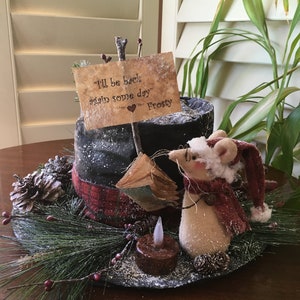 May include: A whimsical holiday centerpiece featuring a mouse figurine wearing a Santa hat and scarf. The arrangement includes a black top hat, a sign that reads "I'll be back again some day" and a faux candle, all set on a bed of pine and berries.