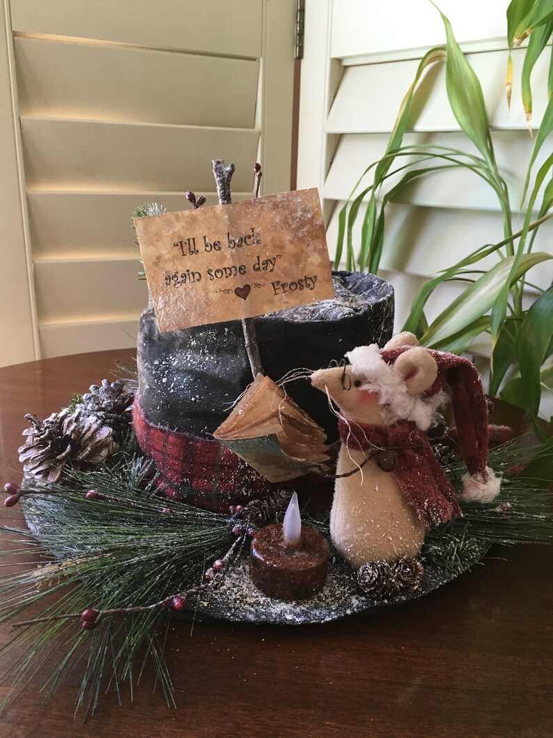 May include: A whimsical holiday centerpiece featuring a mouse figurine wearing a Santa hat and scarf. The arrangement includes a black top hat, a sign that reads "I'll be back again some day" and a faux candle, all set on a bed of pine and berries.