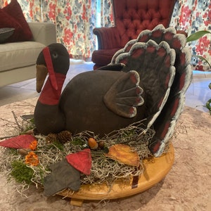 May include: A handcrafted Thanksgiving turkey decoration. The turkey is dark brown with a red wattle and a fan of feathers. It sits on a wooden base surrounded by moss, leaves, and acorns. The turkey is a festive centerpiece.