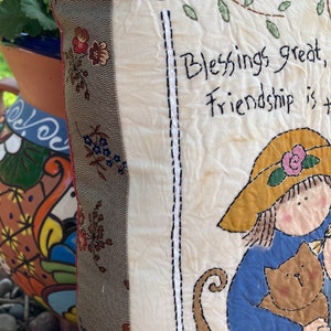 May include: Cream-colored decorative pillow with embroidered text that reads "Blessings great, Friendship is the..." A girl in a hat is holding a brown cat. The pillow has a floral patterned border.