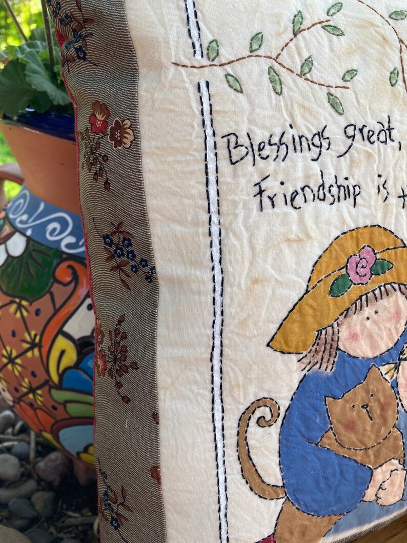 May include: Cream-colored decorative pillow with embroidered text that reads "Blessings great, Friendship is the..." A girl in a hat is holding a brown cat. The pillow has a floral patterned border.