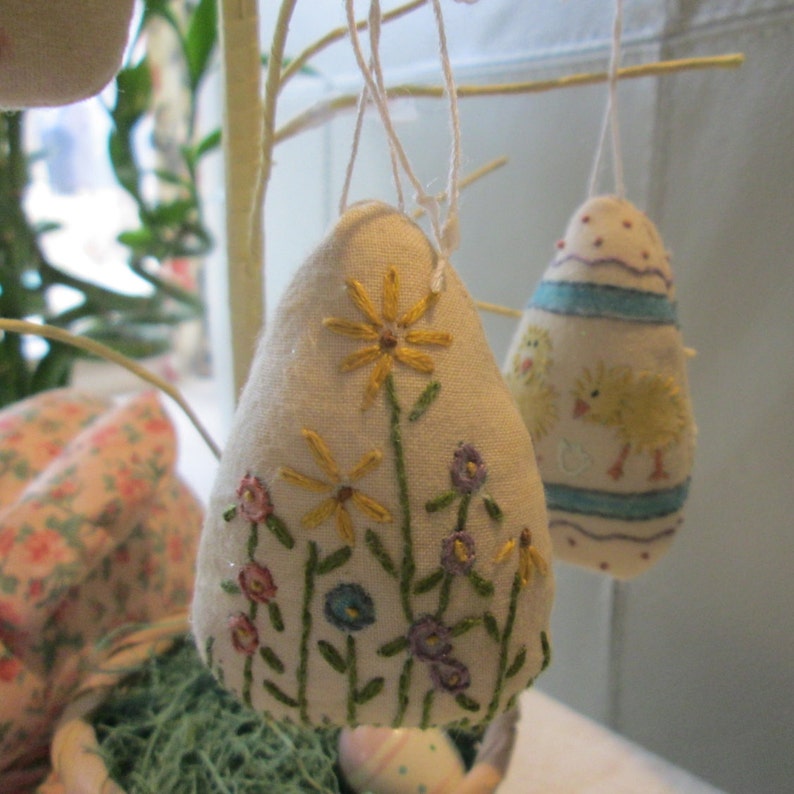 May include: Two hanging Easter egg ornaments. One egg is embroidered with yellow and purple flowers. The other egg has a blue and white striped design with yellow chicks. Both eggs are made of a light-colored fabric.