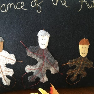 May include: Black felt artwork with the embroidered text "Dance of the Autumn." Four leaf-shaped figures with acorn-shaped heads are dancing. The leaves are white, gray, brown, and green. The figures have smiling faces and are connected by red thread.
