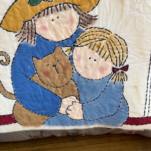 May include: A fabric pillow featuring an appliqued design of two children and a cat. The children are wearing blue shirts and the cat is brown. The pillow has a yellow hat with a pink rose.