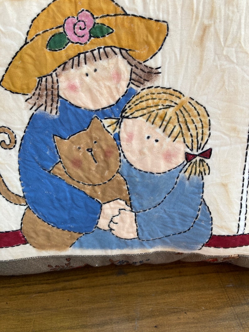 May include: A fabric pillow featuring an appliqued design of two children and a cat. The children are wearing blue shirts and the cat is brown. The pillow has a yellow hat with a pink rose.
