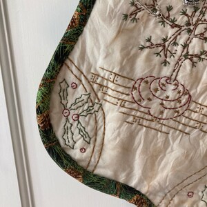 Puede incluir: Calcet&iacute;n navide&ntilde;o color crema con bordados de acebo, notas musicales y un dise&ntilde;o de &aacute;rbol. El calcet&iacute;n est&aacute; adornado con una tela verde con pi&ntilde;as. Un dije plateado de &aacute;rbol de Navidad cuelga del calcet&iacute;n.
