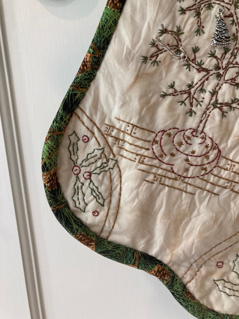 Puede incluir: Calcet&iacute;n navide&ntilde;o color crema con bordados de acebo, notas musicales y un dise&ntilde;o de &aacute;rbol. El calcet&iacute;n est&aacute; adornado con una tela verde con pi&ntilde;as. Un dije plateado de &aacute;rbol de Navidad cuelga del calcet&iacute;n.