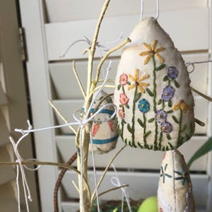 May include: Cream-colored, teardrop-shaped Easter ornaments with embroidered floral designs in yellow, pink, blue, and green hang from a decorative branch. The ornaments are suspended by white string, creating a festive display.