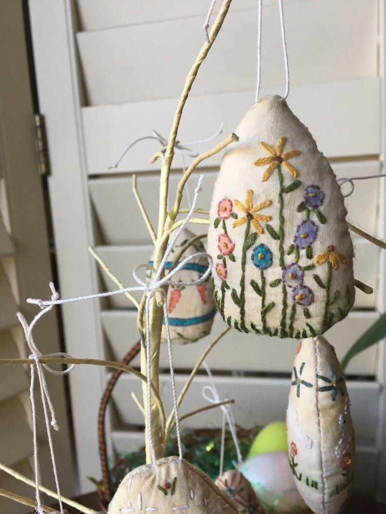 May include: Cream-colored, teardrop-shaped Easter ornaments with embroidered floral designs in yellow, pink, blue, and green hang from a decorative branch. The ornaments are suspended by white string, creating a festive display.