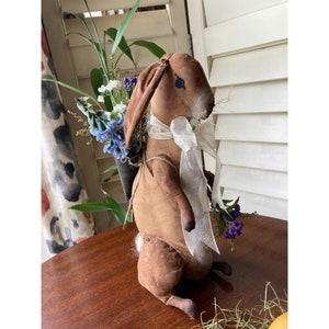 May include: A brown fabric bunny figurine with long ears, holding a small basket of blue and white flowers. The bunny has a white ribbon around its neck and a fluffy white tail. The figurine is on a wooden surface.