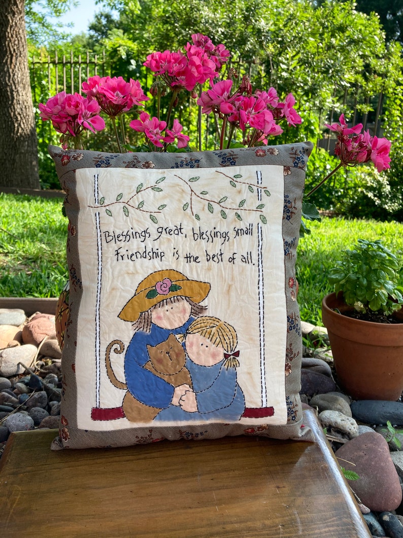 May include: A decorative pillow with a quilted design. The pillow features an illustration of two figures hugging a cat, with the text "Blessings great, blessings small, Friendship is the best of all." The pillow has a floral border and is set against a backdrop of pink flowers.