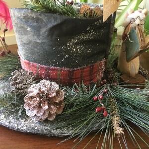 May include: A decorative top hat with a black crown and a red plaid band. The hat is decorated with pine needles, pine cones, and red berries, creating a festive holiday theme. A small figurine of a mouse wearing glasses and holding a book is next to the hat.