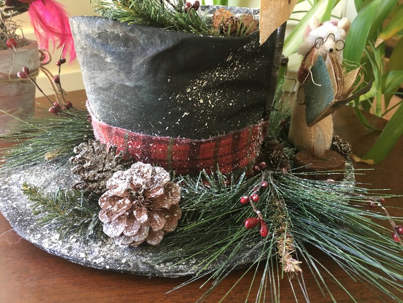 May include: A decorative top hat with a black crown and a red plaid band. The hat is decorated with pine needles, pine cones, and red berries, creating a festive holiday theme. A small figurine of a mouse wearing glasses and holding a book is next to the hat.