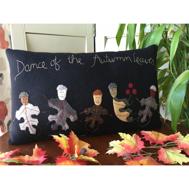May include: A rectangular black pillow with the embroidered phrase "Dance of the Autumn Leaves." The pillow features appliqued leaf figures in various colors, with smiling faces and hats. Decorative fall leaves are scattered in front.