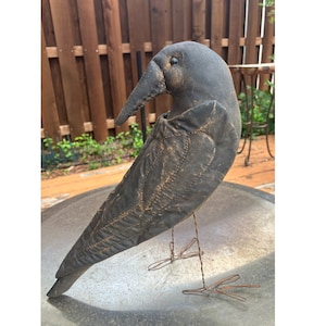 May include: A handcrafted, dark gray fabric crow sculpture with a long beak and wire legs. The crow has stitched details and a slightly distressed appearance, suggesting a rustic or vintage aesthetic. It is perched on a metal surface.