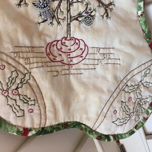 Puede incluir: Falda de &aacute;rbol de Navidad color crema con detalles bordados. Presenta un dise&ntilde;o de &aacute;rbol con base roja y detalles de estrellas, notas musicales y acentos de hojas de acebo. El borde es verde con patrones de bayas rojas. Incluye adornos plateados de &aacute;ngel y copo de nieve.