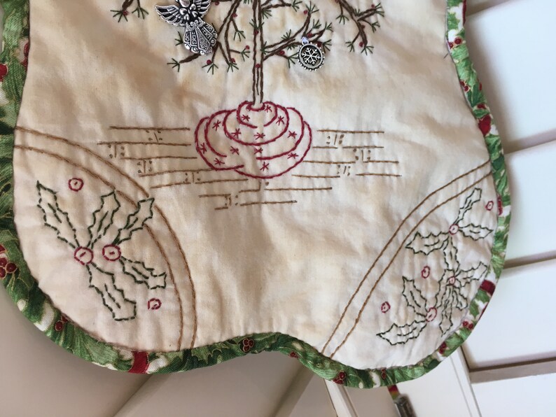 Puede incluir: Falda de &aacute;rbol de Navidad color crema con detalles bordados. Presenta un dise&ntilde;o de &aacute;rbol con base roja y detalles de estrellas, notas musicales y acentos de hojas de acebo. El borde es verde con patrones de bayas rojas. Incluye adornos plateados de &aacute;ngel y copo de nieve.