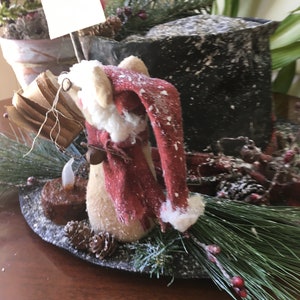 May include: A festive Christmas decoration featuring a Santa Claus figurine with a red hat and scarf, surrounded by pine needles, pine cones, and a small candle. The arrangement is set on a dark, round base, creating a charming holiday display.