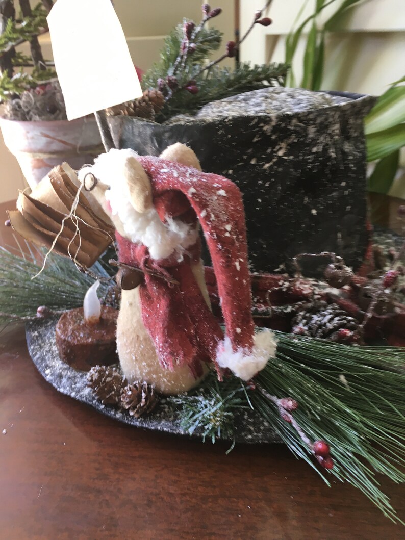 May include: A festive Christmas decoration featuring a Santa Claus figurine with a red hat and scarf, surrounded by pine needles, pine cones, and a small candle. The arrangement is set on a dark, round base, creating a charming holiday display.