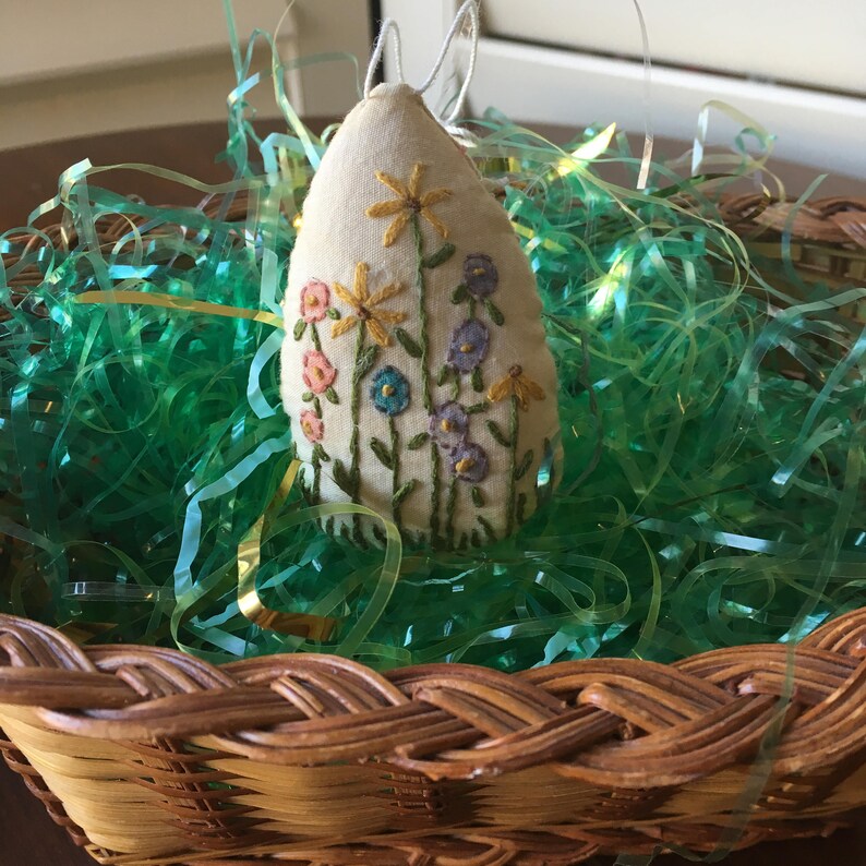 May include: An Easter egg-shaped ornament made of cream-colored fabric, embroidered with colorful flowers. The ornament has a loop for hanging and is nestled in a woven basket filled with green and gold Easter grass. A charming spring decoration.