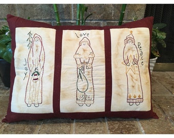 May include: Rectangular decorative pillow with three panels featuring embroidered Santa Claus figures. The pillow has a burgundy border and vertical stripes. The words "joy", "love", and "peace" are embroidered on the pillow.