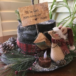 May include: A whimsical winter decoration featuring a mouse wearing a Santa hat and scarf. A sign reads "I'll be back again some day" with a heart. The arrangement includes a faux candle, pine needles, and a plaid accent.