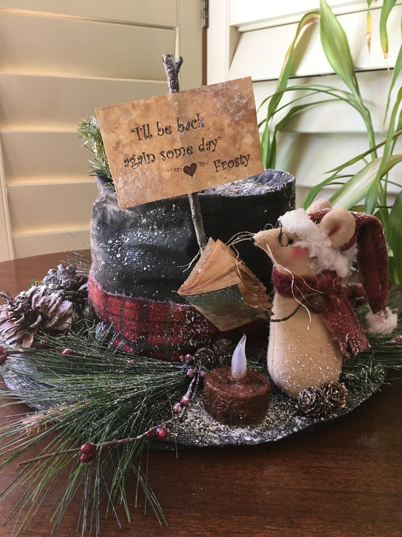 May include: A whimsical winter decoration featuring a mouse wearing a Santa hat and scarf. A sign reads "I'll be back again some day" with a heart. The arrangement includes a faux candle, pine needles, and a plaid accent.