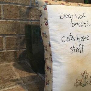 May include: Square decorative cushion with the text "Dogs have owners... Cats have staff" embroidered in black thread. The cushion has a floral patterned border and embroidered flowers. The background is a brick wall and a green pot.