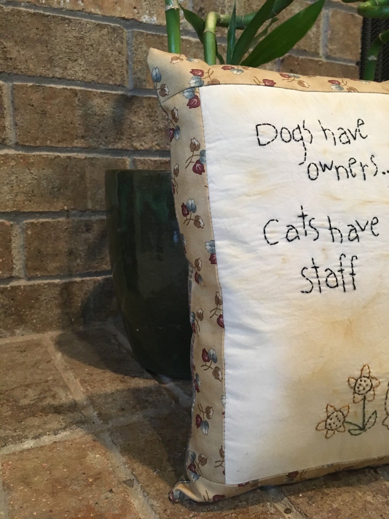 May include: Square decorative cushion with the text "Dogs have owners... Cats have staff" embroidered in black thread. The cushion has a floral patterned border and embroidered flowers. The background is a brick wall and a green pot.