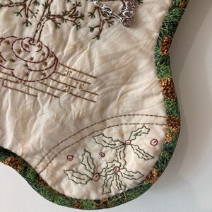Puede incluir: Calcet&iacute;n navide&ntilde;o color crema con dise&ntilde;os bordados de &aacute;rboles y acebo. Amuletos plateados de un mu&ntilde;eco de nieve, un calcet&iacute;n y un esqu&iacute; adornan el calcet&iacute;n. El borde est&aacute; adornado con un patr&oacute;n de pi&ntilde;as verdes y marrones.
