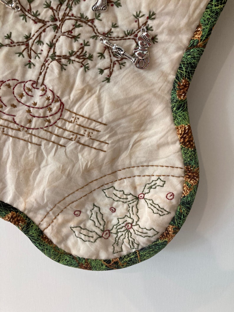 Puede incluir: Calcet&iacute;n navide&ntilde;o color crema con dise&ntilde;os bordados de &aacute;rboles y acebo. Amuletos plateados de un mu&ntilde;eco de nieve, un calcet&iacute;n y un esqu&iacute; adornan el calcet&iacute;n. El borde est&aacute; adornado con un patr&oacute;n de pi&ntilde;as verdes y marrones.