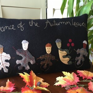 May include: Black rectangular pillow with the embroidered text "Dance of the Autumn Leaves." The pillow features appliqued leaf figures with smiling faces. Colorful fall leaves are arranged in the foreground.