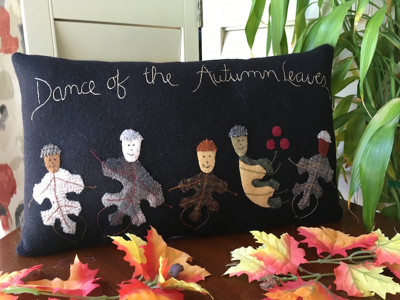 May include: Black rectangular pillow with the embroidered text "Dance of the Autumn Leaves." The pillow features appliqued leaf figures with smiling faces. Colorful fall leaves are arranged in the foreground.