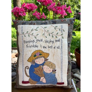 May include: Square decorative pillow with a stitched design. The pillow features a central panel with the text "Blessings great, blessings small, Friendship is the best of all." The panel depicts two children and a cat. The pillow has a floral border.