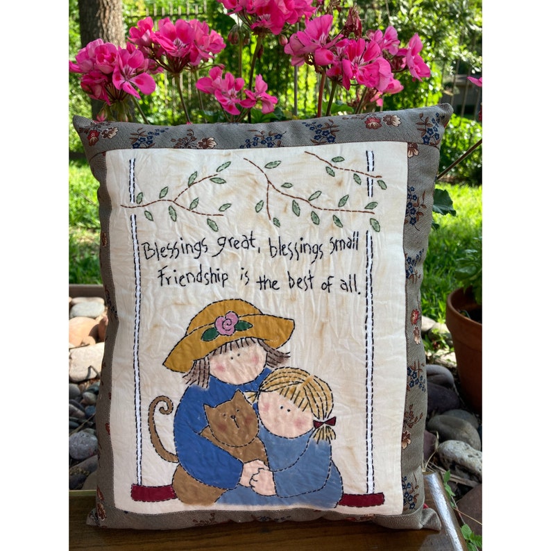 May include: Square decorative pillow with a stitched design. The pillow features a central panel with the text "Blessings great, blessings small, Friendship is the best of all." The panel depicts two children and a cat. The pillow has a floral border.